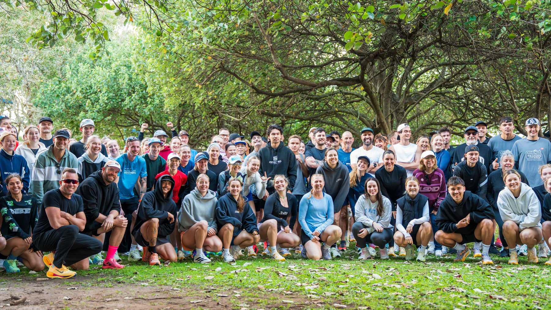 A crowd of around a hundred runners crouched together under trees after a shakeout run, most of them smiling at the camera.