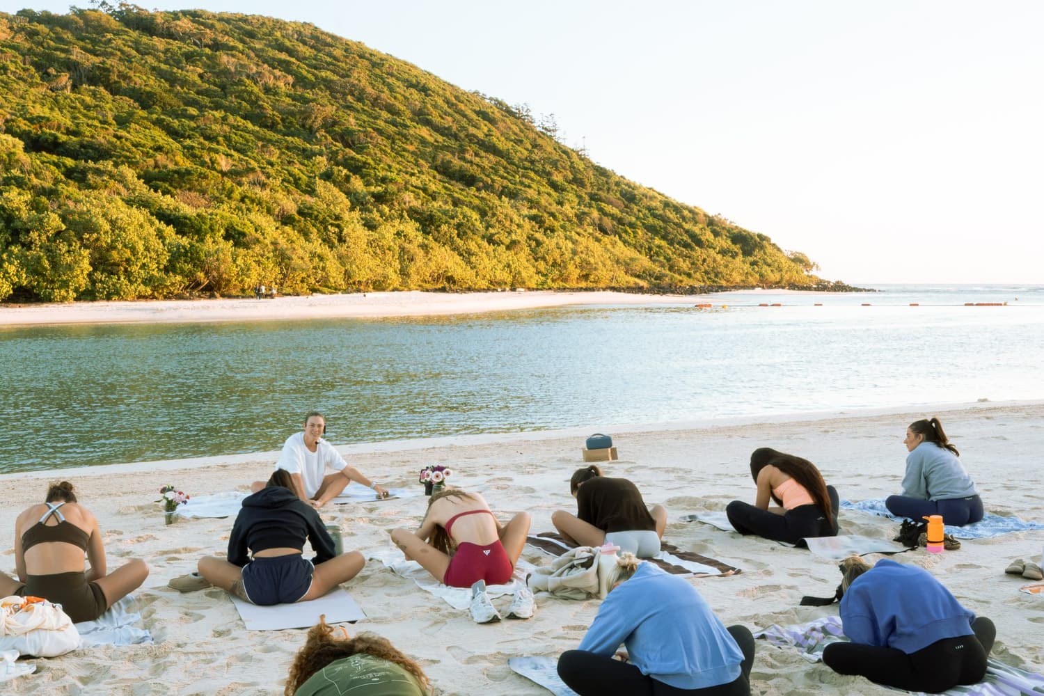 A beach mobility class seated on mats facing the water and the green headland.